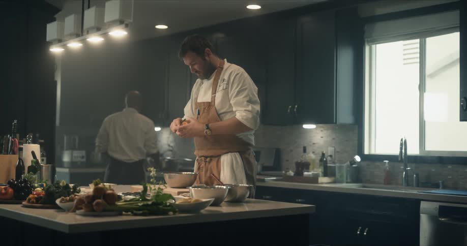Man peels an apple at kitchen island as another man works near stove