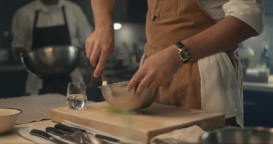 A man wearing an apron is stirring something in a silver bowl and another man brings him a bigger silver bowl