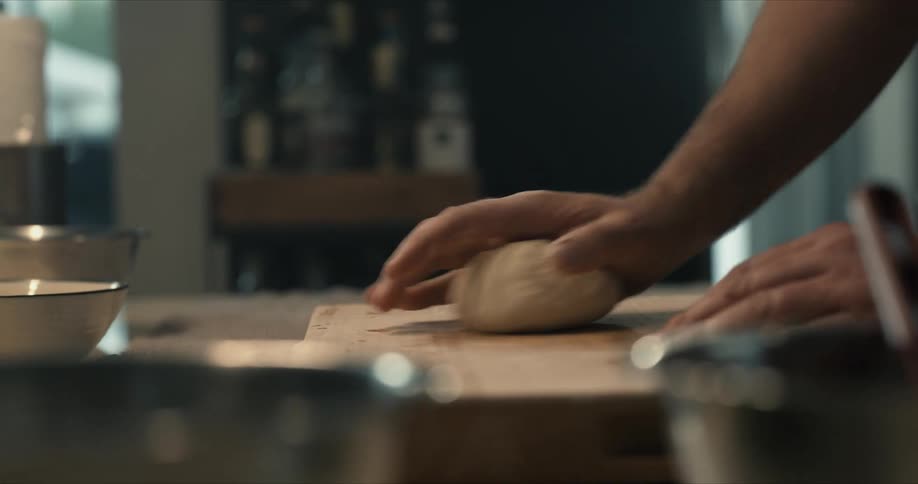 A man kneads dough on a wooden cutting board in a kitchen during the day