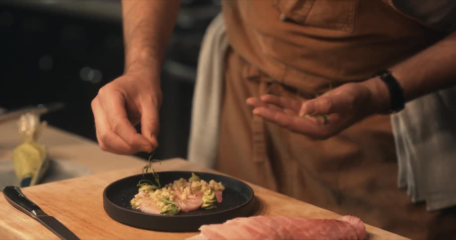 Chef garnishes a dish with herbs over a black dish in the kitchen
