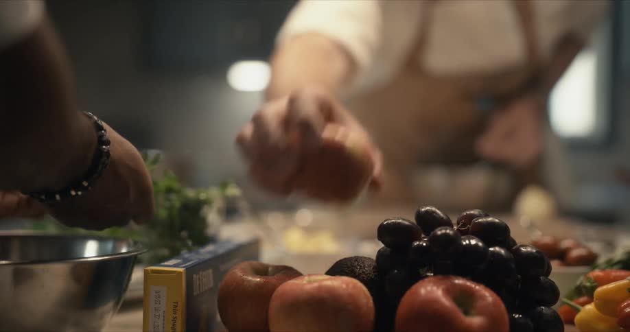 A chef reaches across and picks up an apple near another chef placing parsley sprigs in a bowl