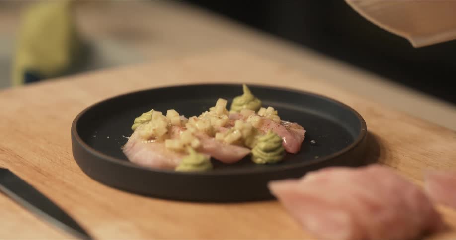 Chef garnishes a raw fish dish on a black plate over the table