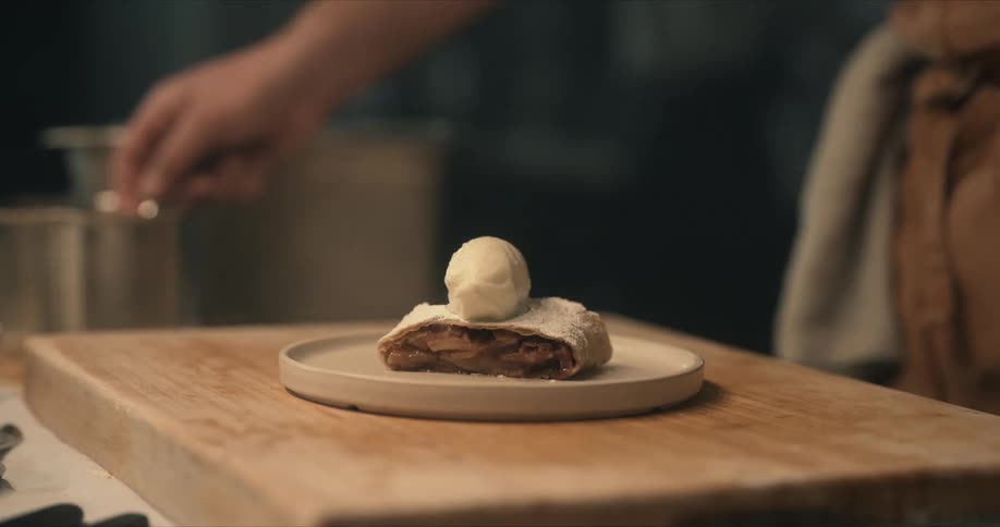 A man in a kitchen puts a small scoop of ice cream on top of a piece of apple strudel sitting on a plate and hands it to another man