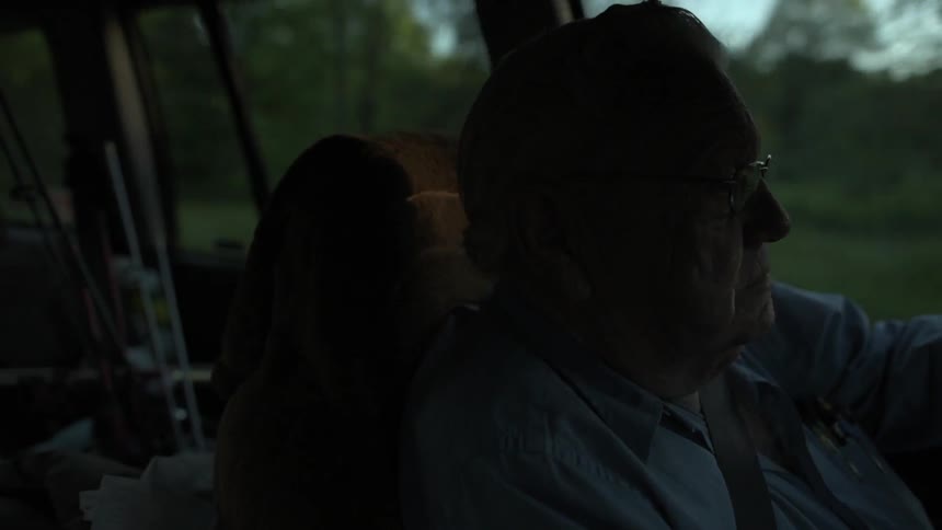 A man is riding in a vehicle past green fields with trees