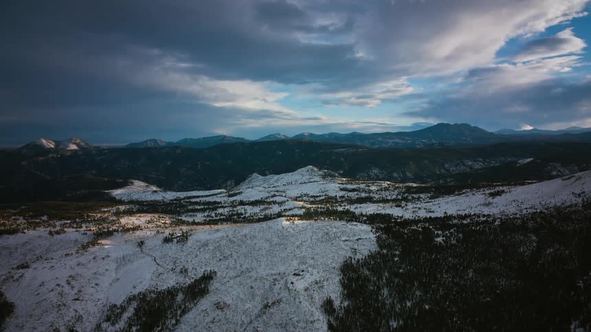 Clouds move over snow covered forested mountains and there are mountains in the distance