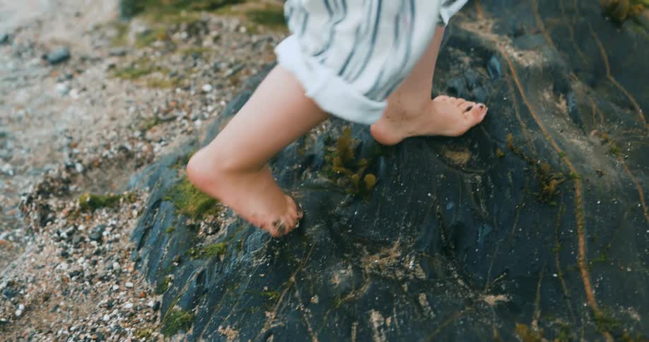 A woman walks alongside a small child walking barefoot on stones in a creek