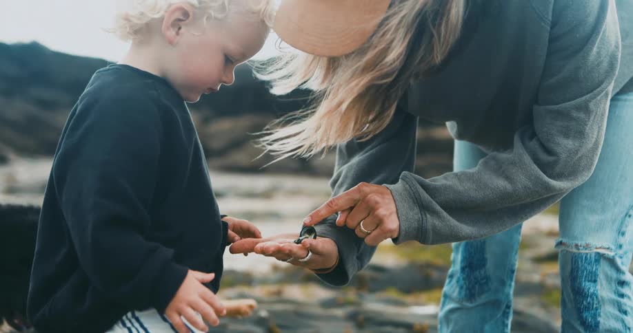 A woman shows her son rocks in her hand as they stand on the beach