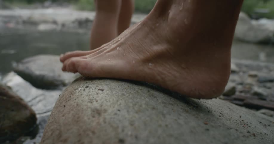 A pair of people walk barefoot across stones spanning a stream during the day