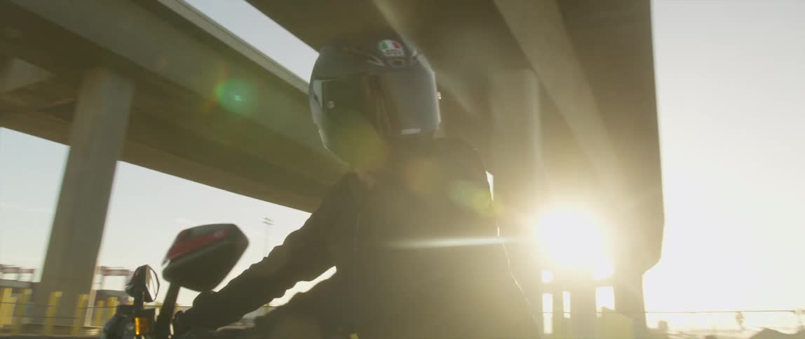 A motorcyclist looks around at their surroundings while sitting on a motorcycle beneath a bridge during the day