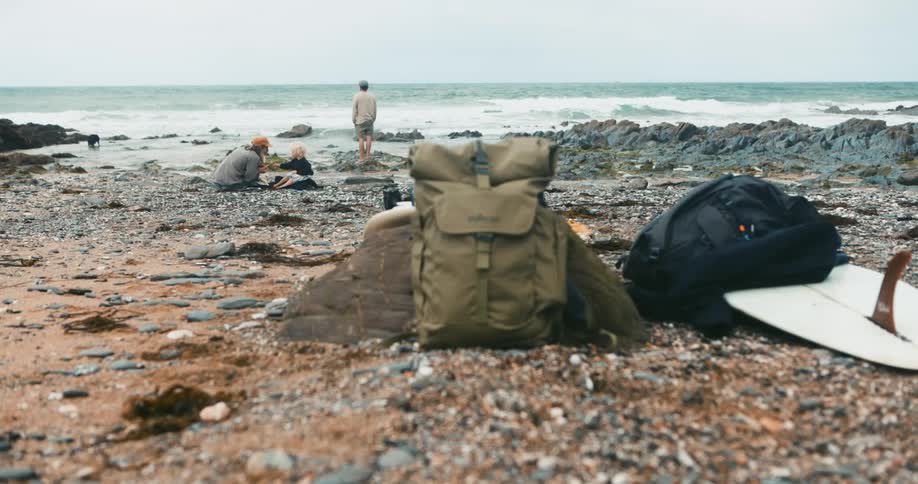 Father hops onto rocks near ocean as mother sits with childand their gear including a surfboard sits on rocky ground further from the water
