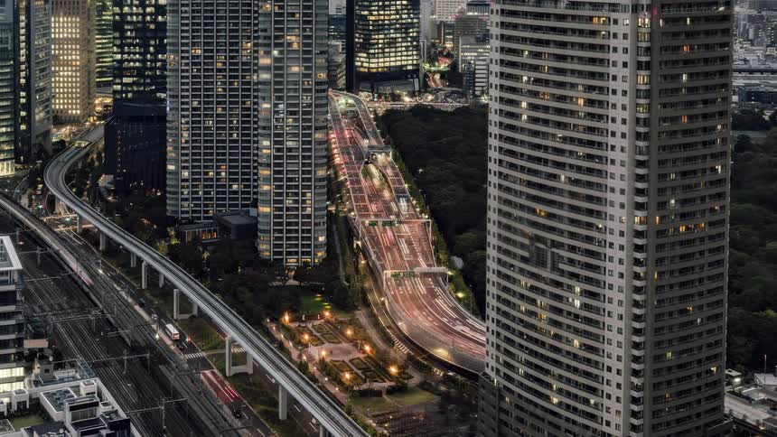 Traffic moves on busy a highway and train tracks around a big city with towers and skyscrapers that light up when it gets dark outside