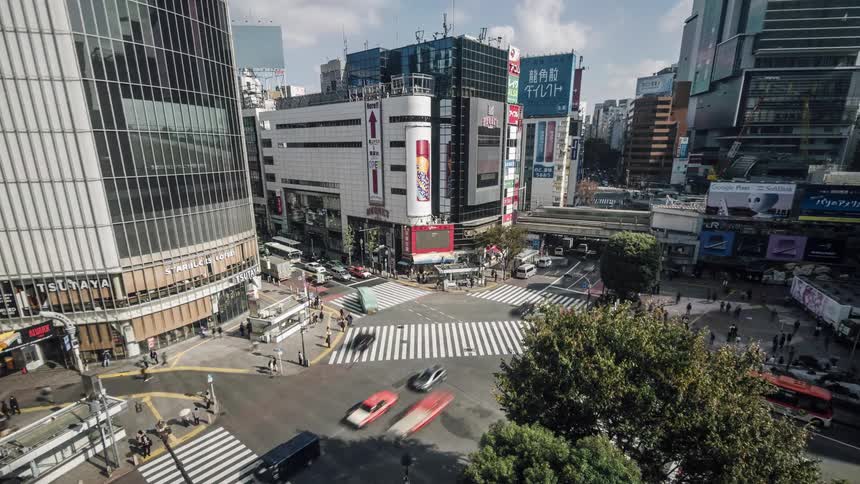 People are walking on a downtown intersection and advertisements are on big screens on buildings as cars and buses move on the streets and a train crosses a bridge