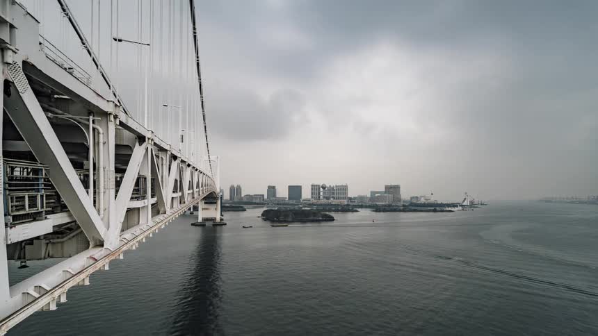 Boats are sailing under a bridge as a train passes by and big city buildings can be seen in the distance under a cloudy sky