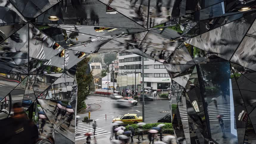 Pedestrians holding umbrellas and traffic on the street reflect in the mirrors inside a structure on a rainy day