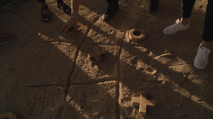 A small group of teenagers are playing tic tac toe in the dirt with wooden pieces