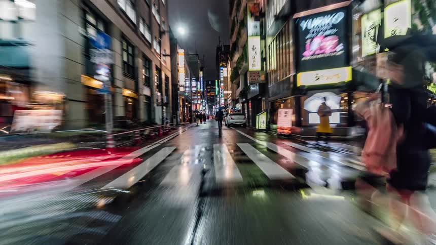 Pedestrians holding umbrellas walk towards an intersection as traffic travels on the street in the city at night