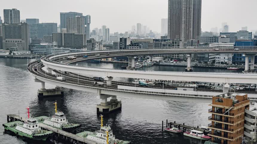 A train goes around a curve over a river and there is traffic on the highway below and big city buildings in the distance