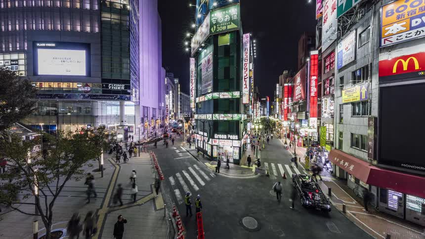 People are walking on a busy downtown street and lights are flashing on buildings and on big screens on buildings as cars pass by