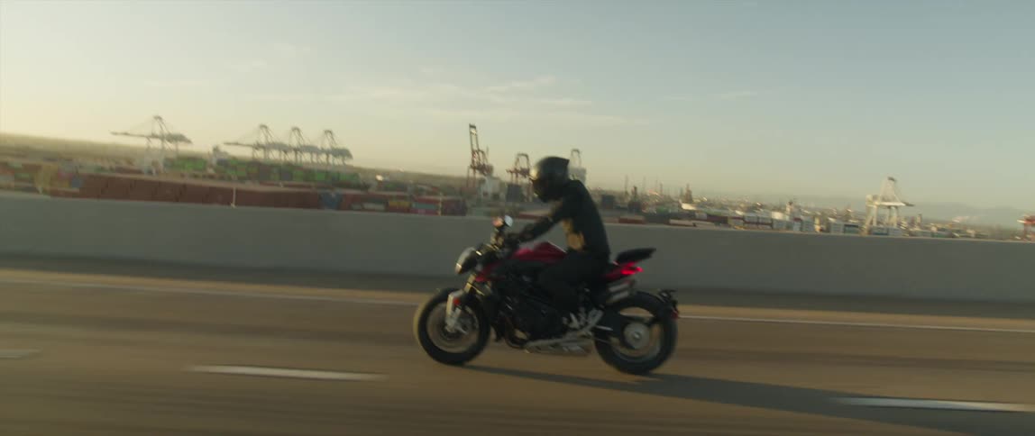 A cyclist wearing a black helmet is riding a motorcycle on a highway and there are construction cranes in the distance