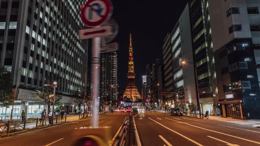 People are waking on a downtown street near tall buildings at night and there is a tower lit up at the end of the street