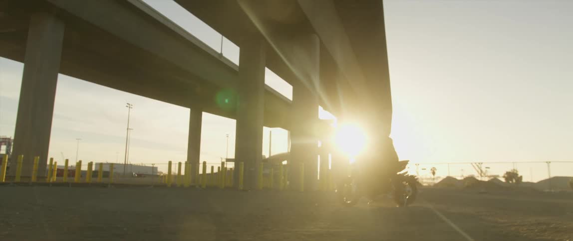 A motorcycle is parked below a highway and someone wearing a black helmet walks to it and gets on as the shines brightly in the distance