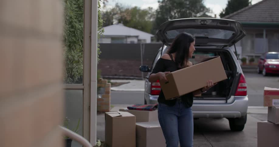 A woman lifts a box from her car and brings it to a man standing in a doorway
