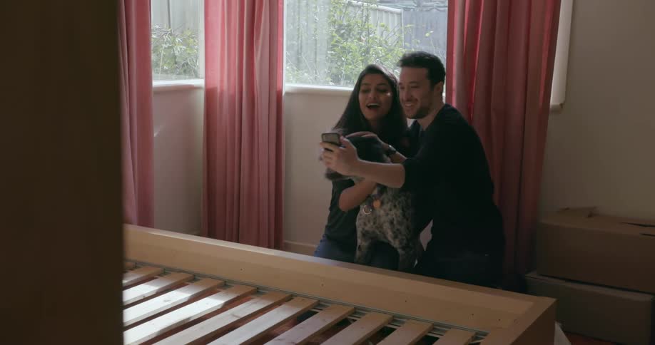 A man and woman play with their dog in a bedroom and take a selfie
