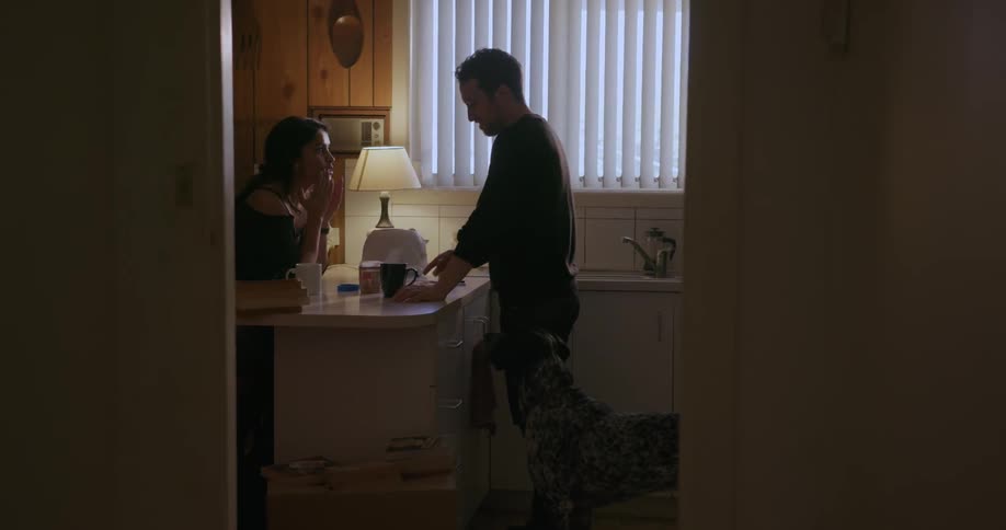 A man and woman are standing at the kitchen counter drinking coffee while their dog looks at them