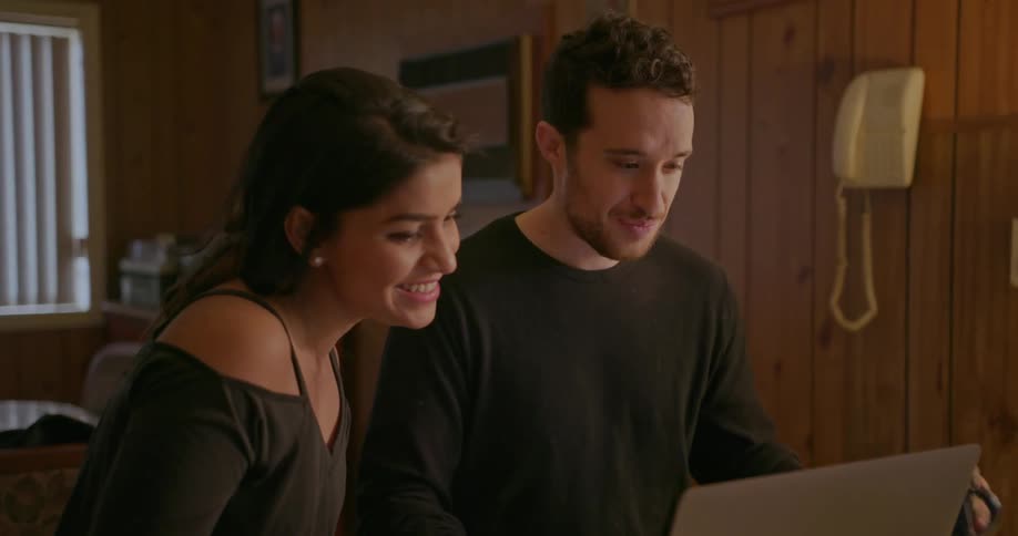 A man and woman sit at a kitchen counter and look at a laptop