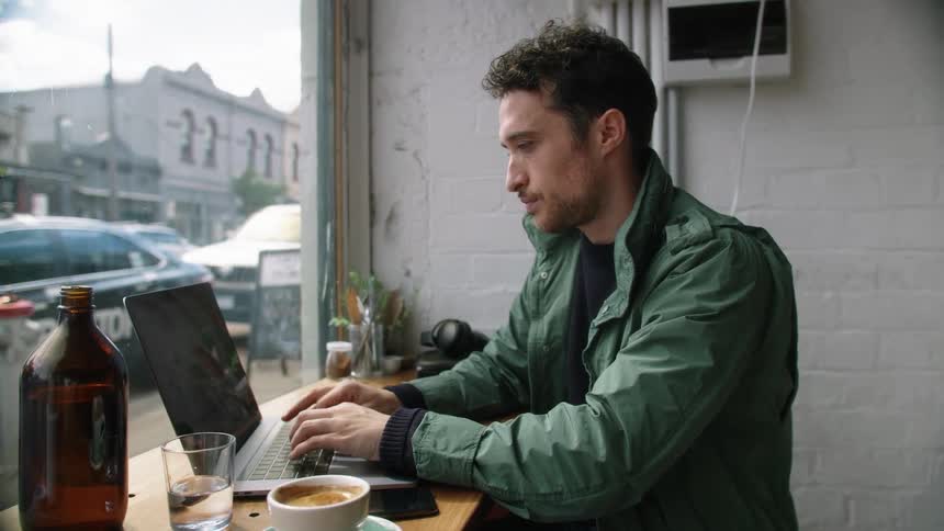 A man is typing on a laptop as he looks out a coffee shop window during the day