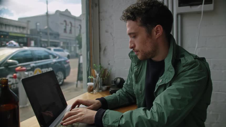 Man sits in a cafe typing on a laptop in a busy city