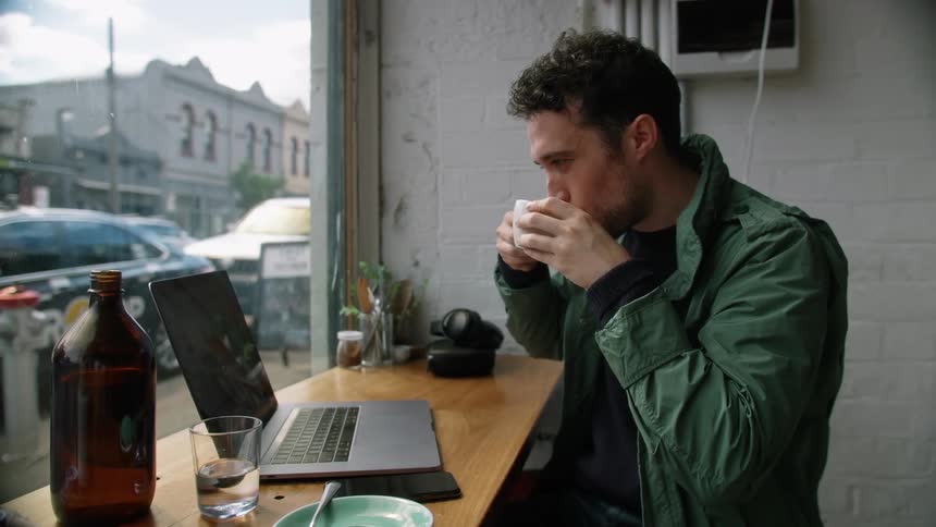 Man sits in a cafe sipping from a mug and looking at a laptop