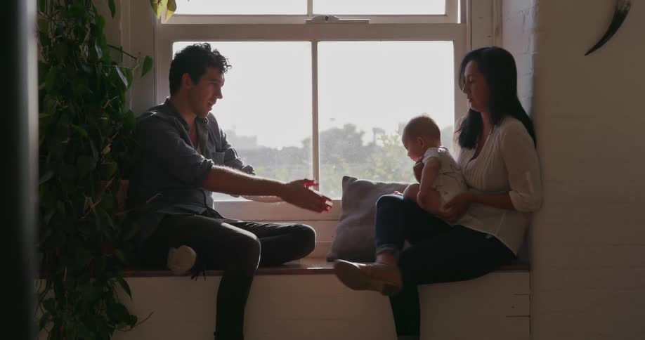 A mother and father are sitting in a window with their baby in daylight