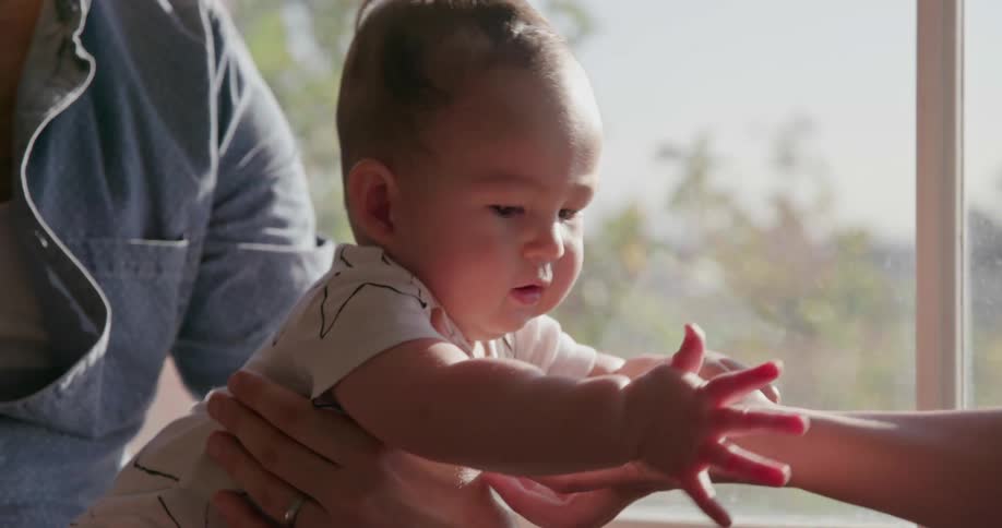 A baby holds hands with a person by the window