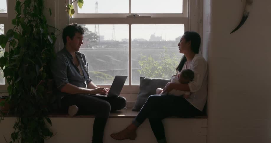 A man is working at a computer while sitting with a woman and baby in a window