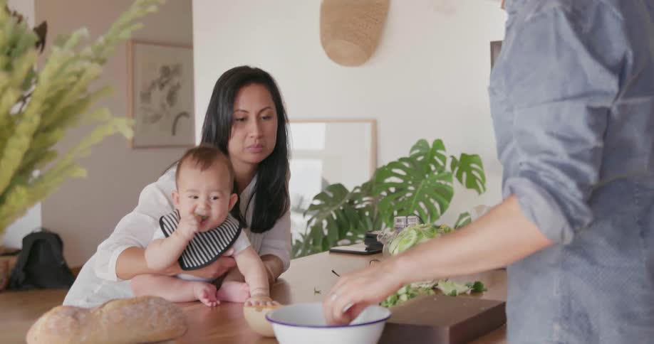 A woman is sitting at the table holding a baby while a man is cutting veggies