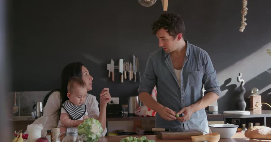 Woman holds a child that is playing with a head of cauliflower and a man talks to the woman while preparing food