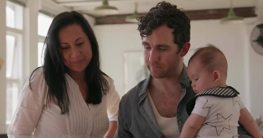 Man and woman looking at a laptop and talking while the man holds an infant indoors