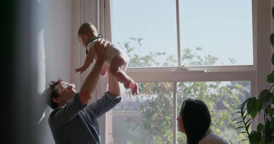 Man and woman sitting next to a window and a man raises an infant into the air and then brings back down