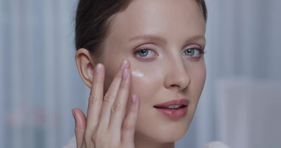 A model looks into the camera while rubbing a cream onto a cheek