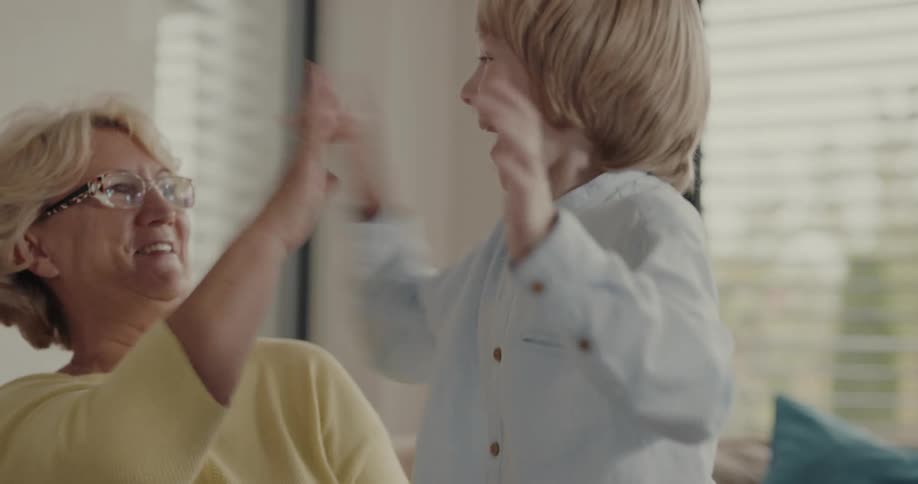A child and his grandmother sit together on a couch looking down at something react with laughter and high five each other and then hug