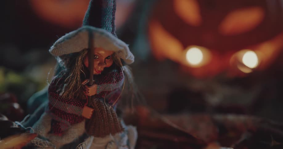 Close up of a small witch toy doll sitting over dry leaves near halloween pumpkins