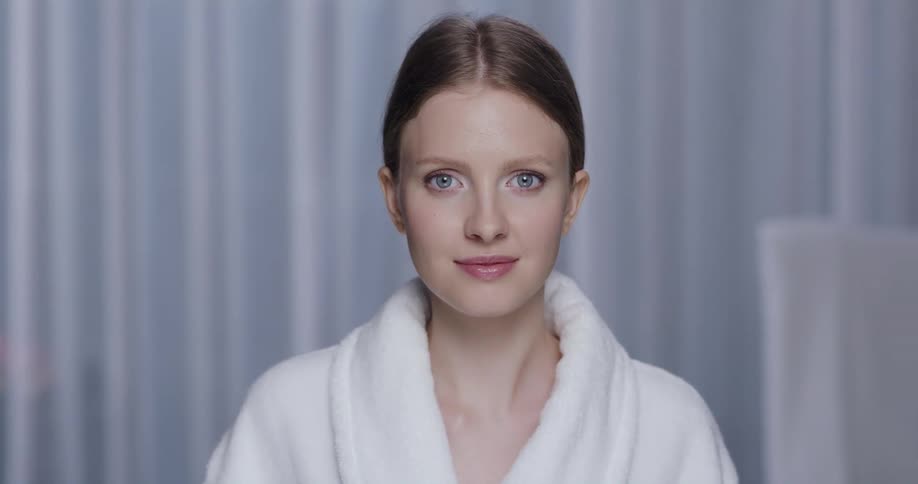 A woman is wearing a white bath robe and there is a white curtain behind her