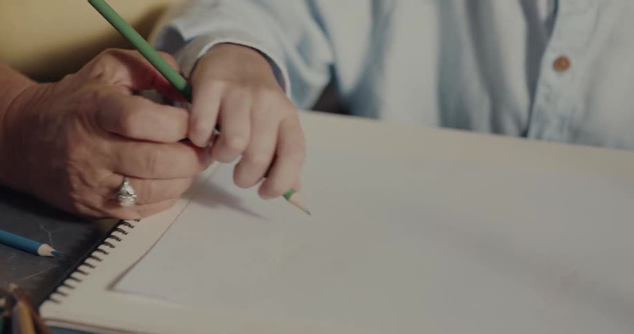 An adult helps a young boy hold a pencil and draw on a notebook