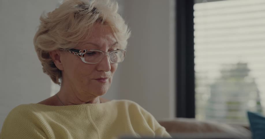 An elderly woman uses a laptop while sitting on a sofa indoors during the day
