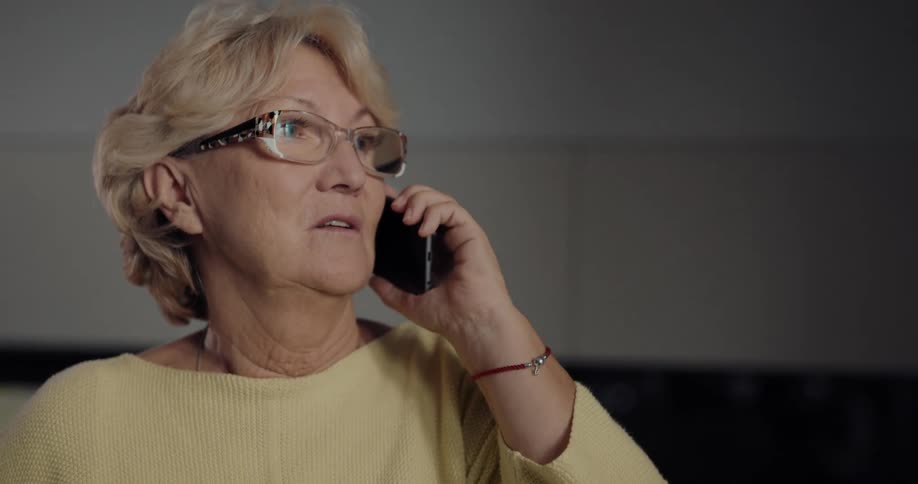 An elderly woman with blond hair and glasses is talking on the phone