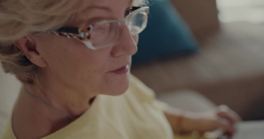 An elderly woman with blond hair and glasses is sitting on a couch looking at a book
