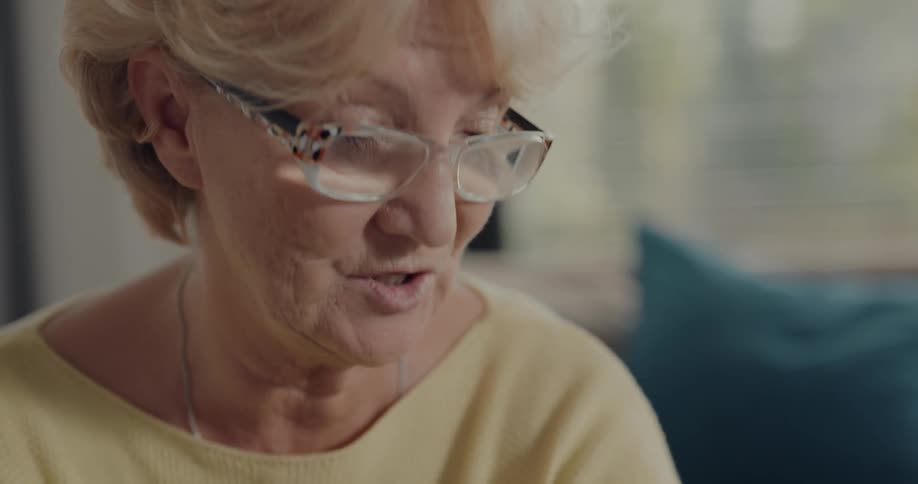 Elderly woman with glasses fair hair and yellow shirt talks and smiles at person sitting on blue couch in living room
