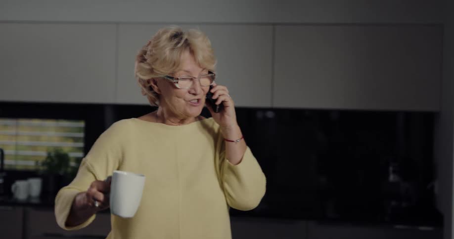 Female senior citizen drinks coffee while talking on the phone in the office