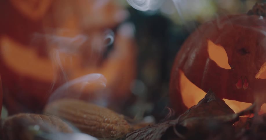 Vapor rises over lit jack o lanterns sitting on ground with dried corn cobs